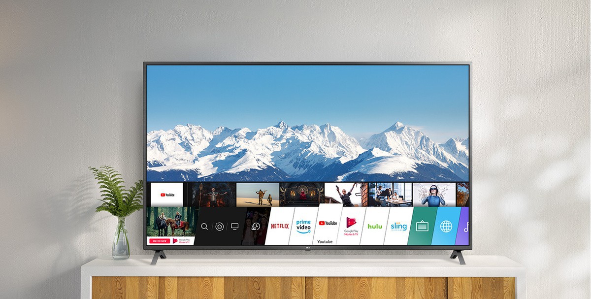 Televizor amplasat pe un suport alb, pe un perete alb. Ecranul TV prezintă ecranul principal cu webOS.