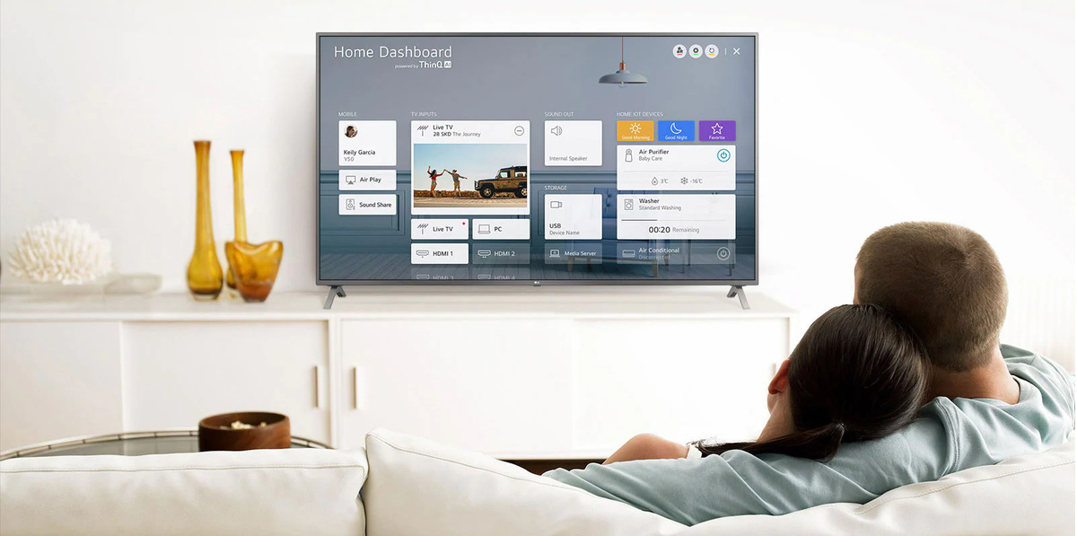 Un bărbat si o femeie care stau pe o canapea în camera de zi, cu Home Dashboard pe ecranul TV
